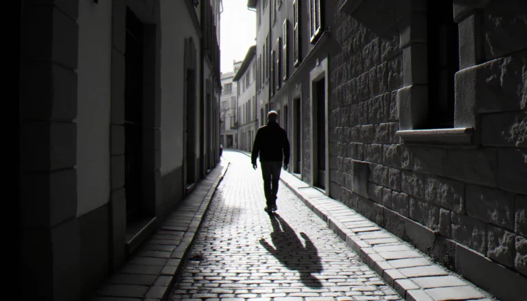 Photographie noir et blanc d'une ruelle pavée avec une silhouette en contre-jour, ombres profondes et lumière rasante sur les murs en pierre